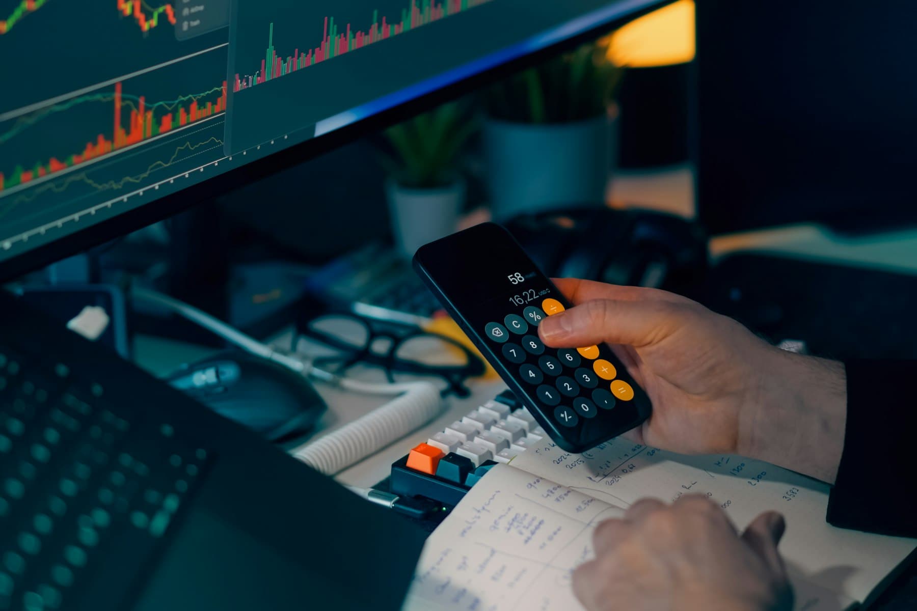 Hand using a calculator at a desk in front of monitors with charts